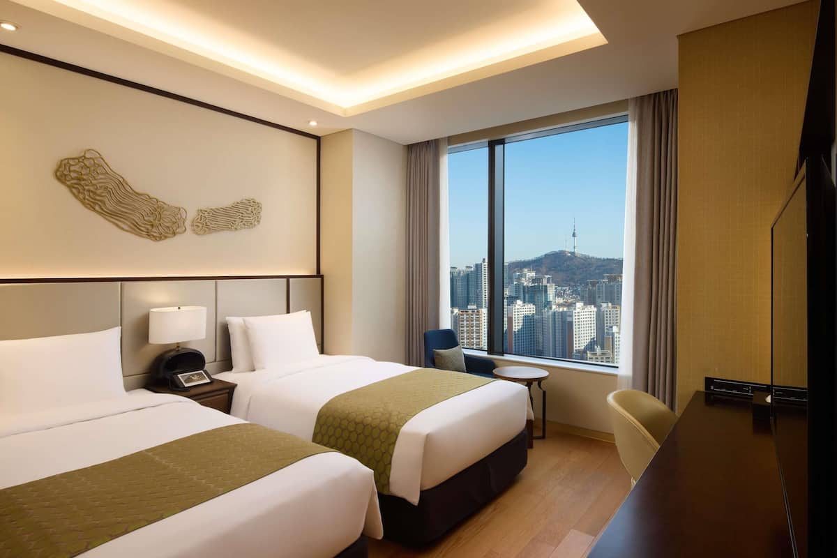 Une chambre d’hôtel avec deux lits, un bureau, une chaise, une télévision et une grande fenêtre donnant sur la ville.