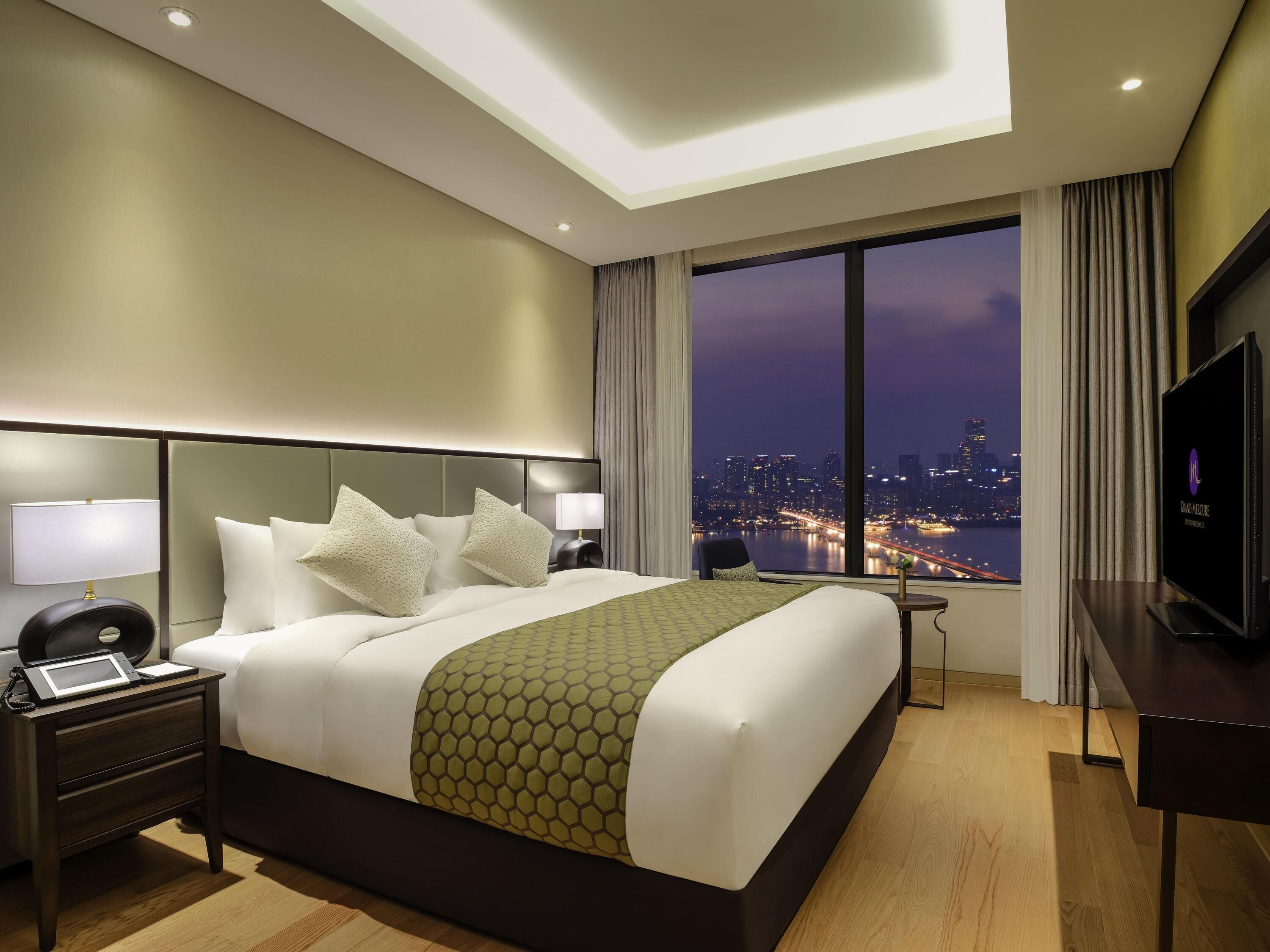 Ein Hotelzimmer mit einem großen Bett, einem Schreibtisch, einem Fernseher und Blick auf die Stadt.