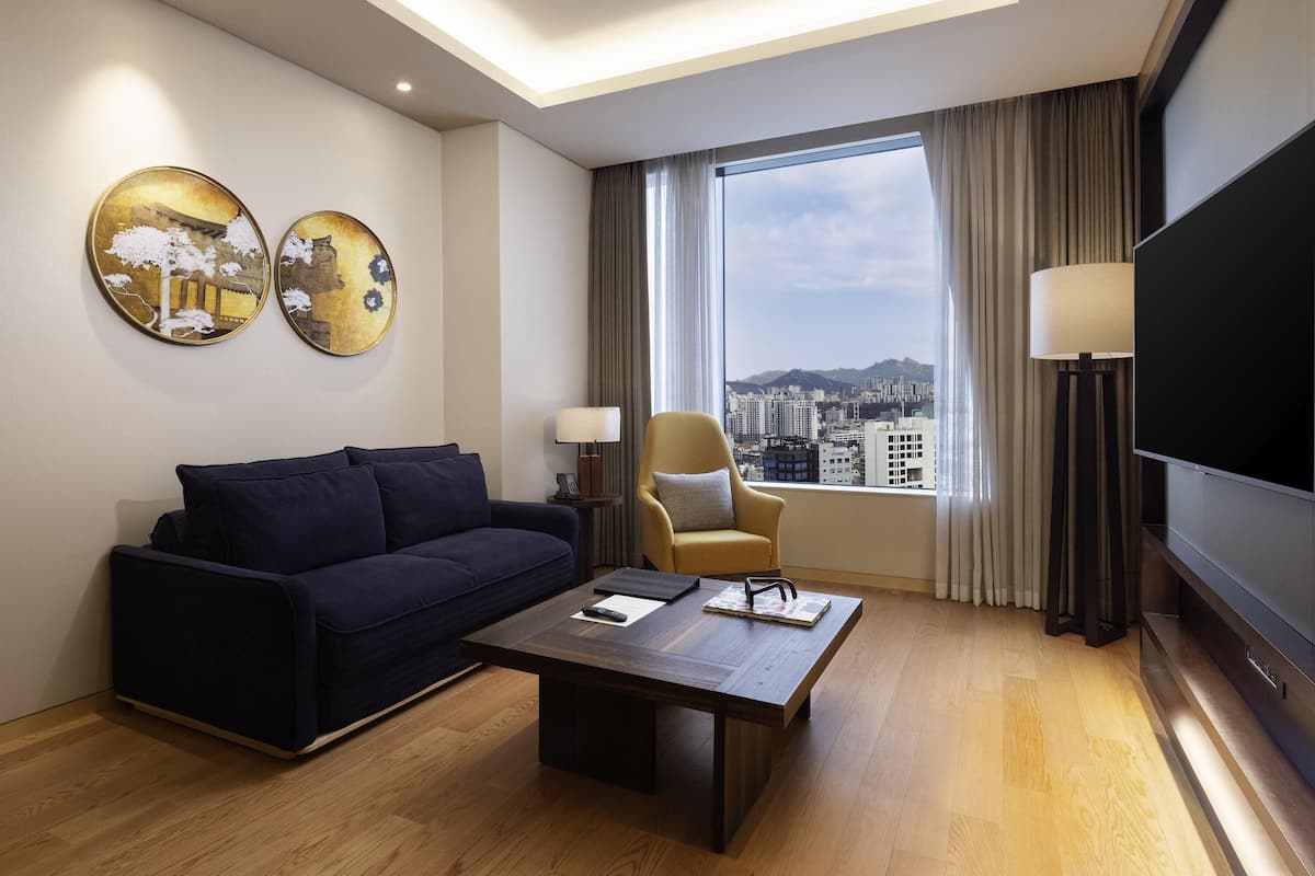 Une chambre d’hôtel moderne avec un canapé bleu foncé, une table basse en bois, un téléviseur à écran plat et une grande fenêtre donnant sur la ville.