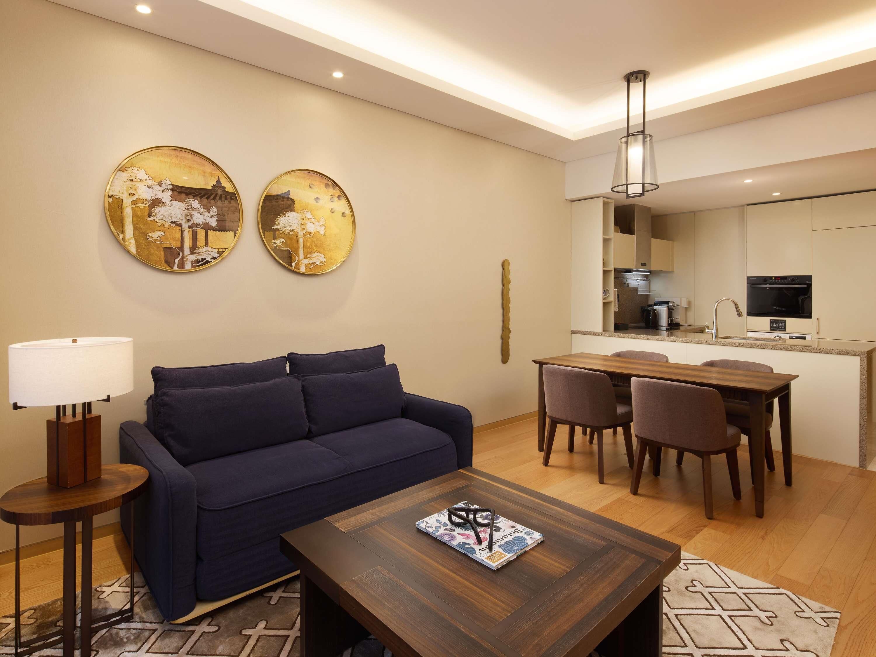 A modern living room with a dark blue sofa, a wooden coffee table with a magazine and glasses, a lamp, and two gold-framed wall art pieces.