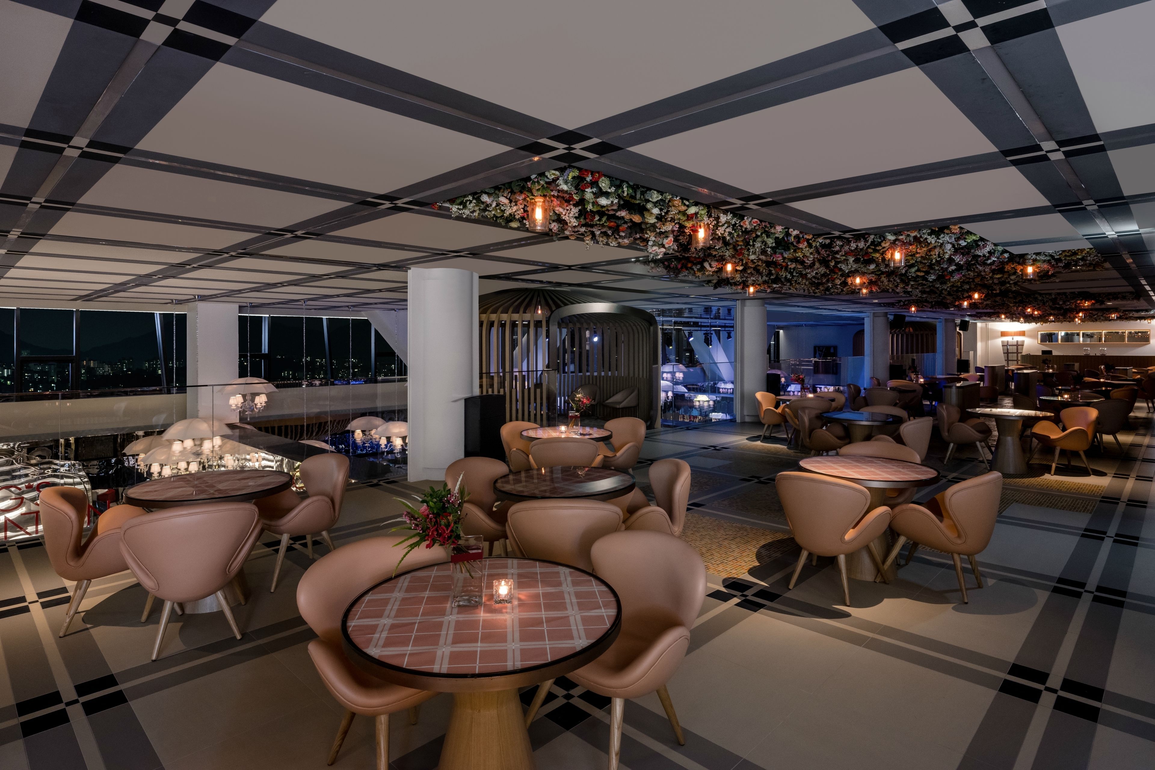 A modern lounge area with round tables and chairs, a checkered ceiling, and a decorative floral arrangement.
