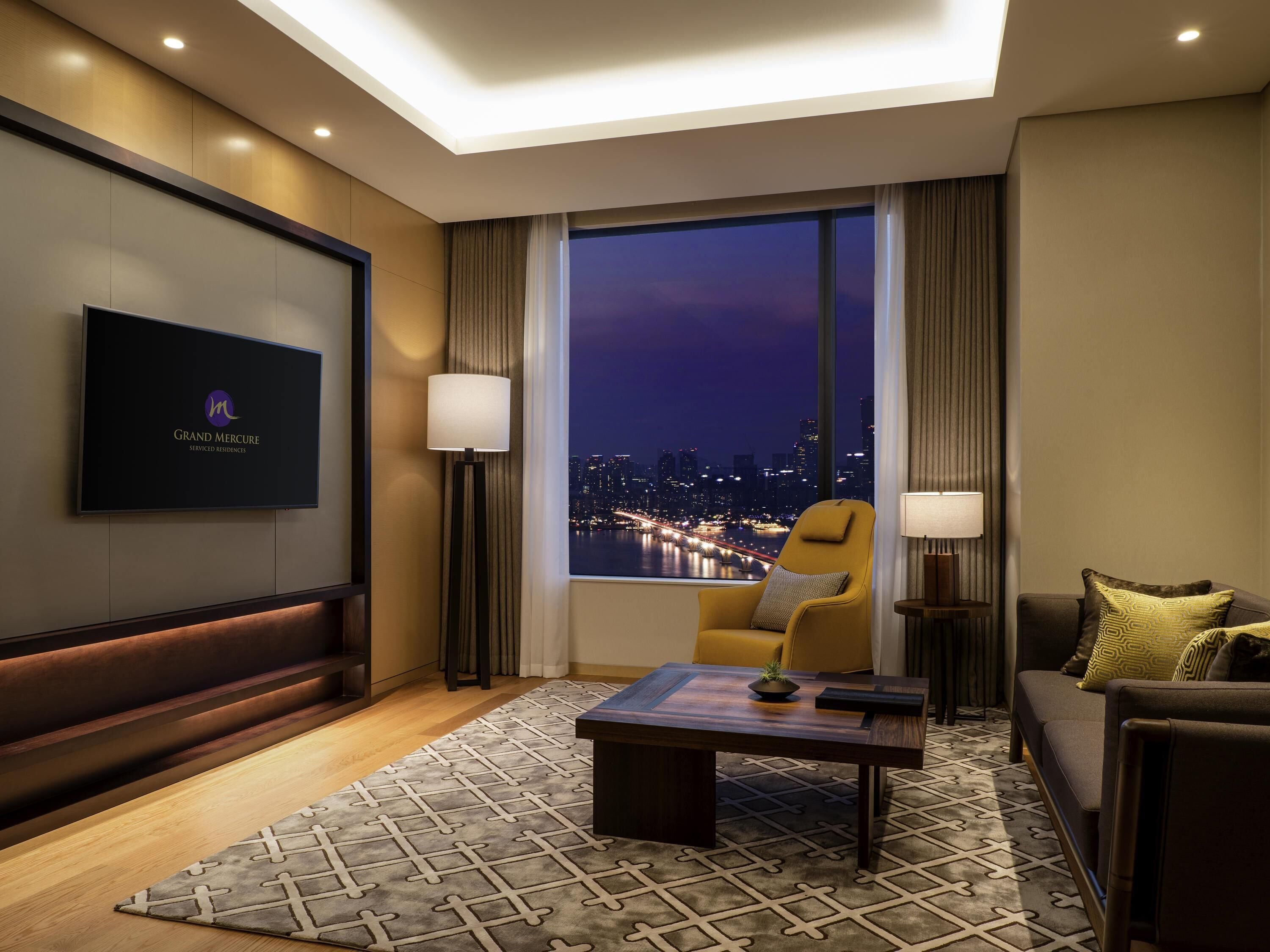 A hotel room with a city view, a flat-screen TV, a sofa, a coffee table, and a patterned rug.