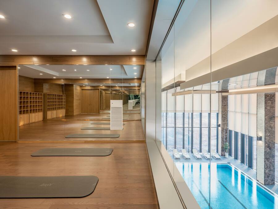 Un gimnasio moderno con pisos de madera, colchonetas de ejercicio y una pared de vidrio con vista a una zona de piscina.