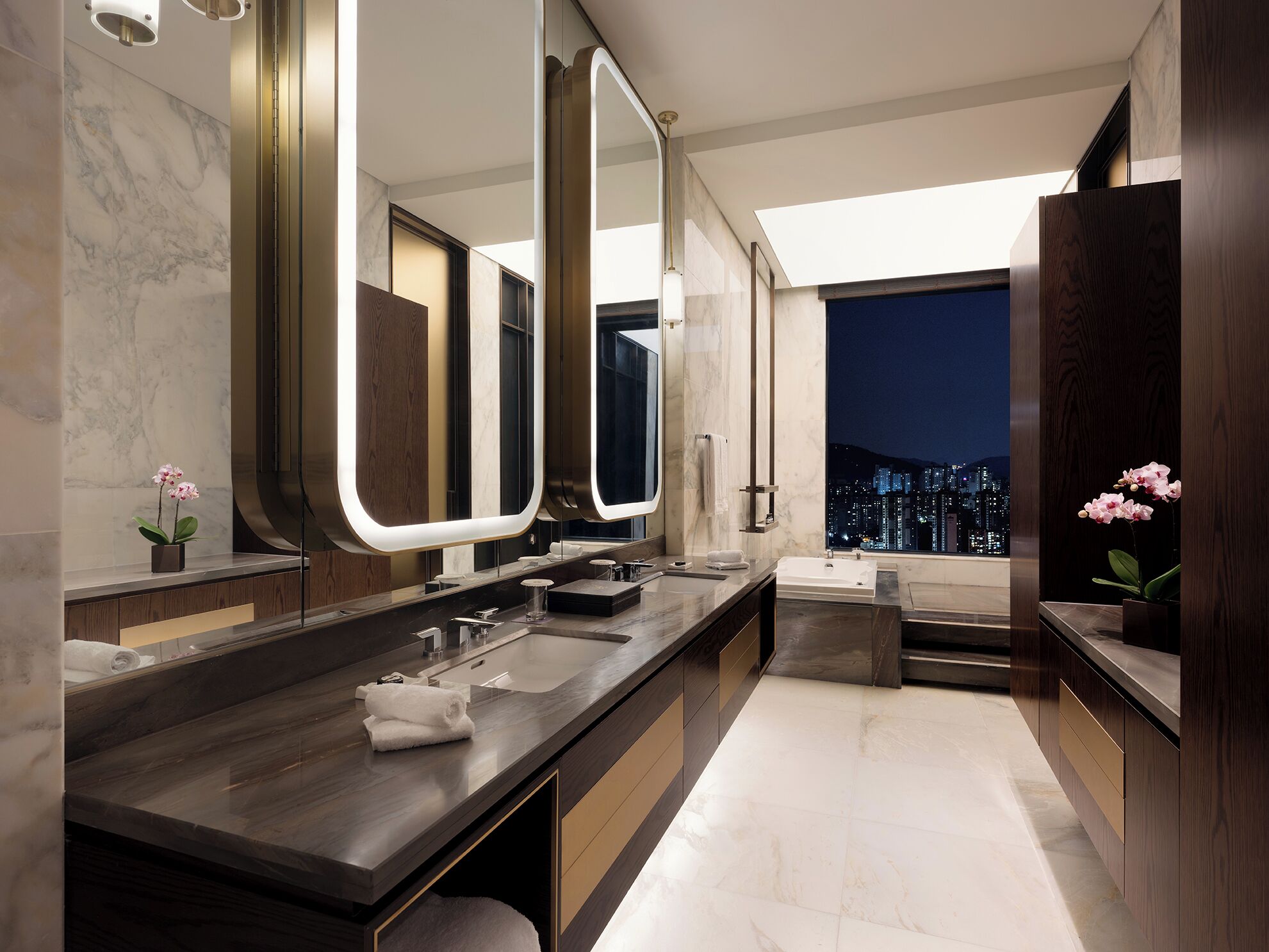 Un baño moderno con un espejo grande, una bañera y vistas a un paisaje urbano nocturno.