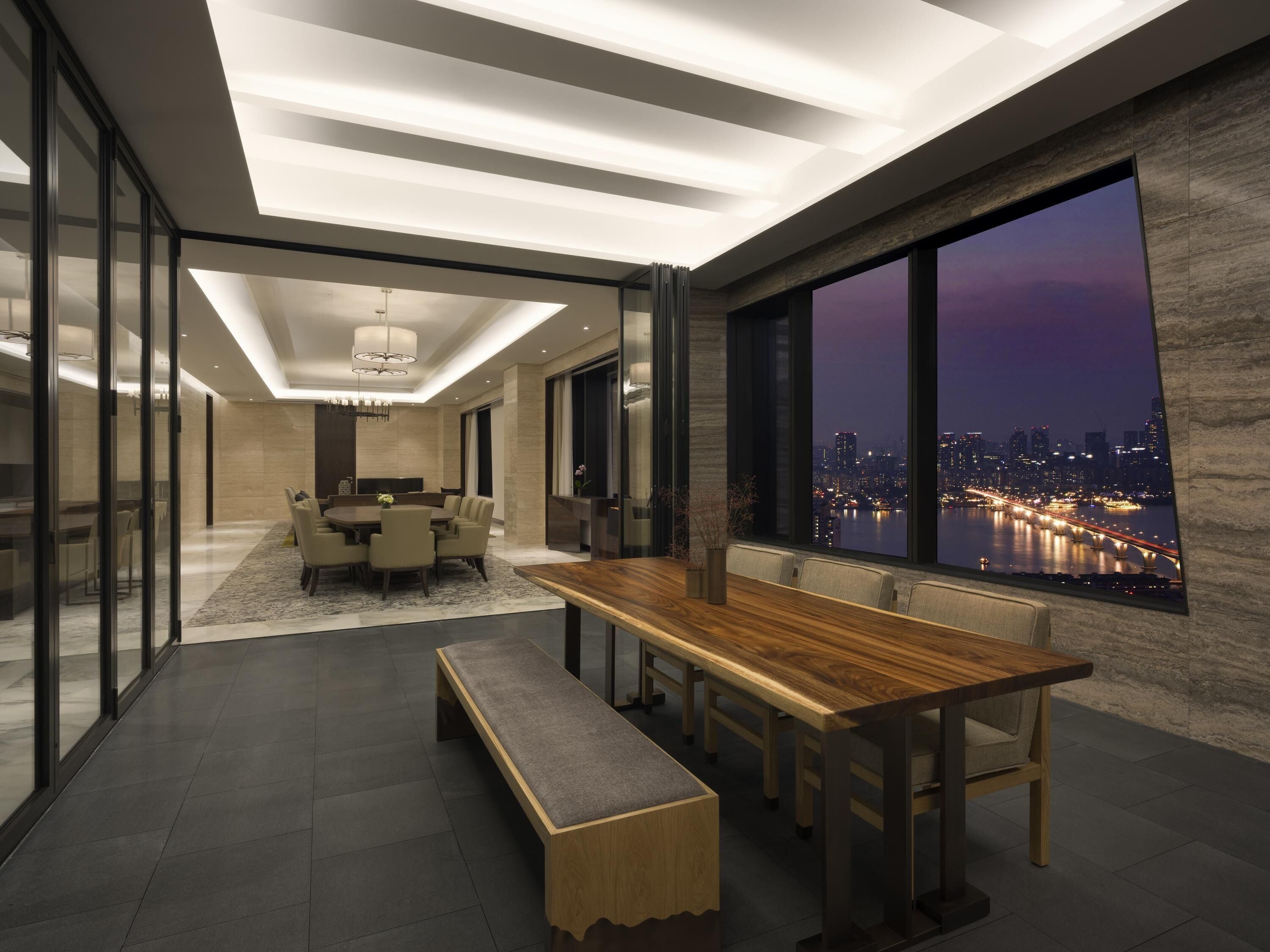 A modern dining area with a long wooden table, benches, and a city view.