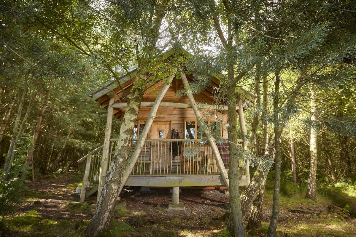 A wooden cabin with a wooden deck and a small porch, surrounded by trees in a forest.