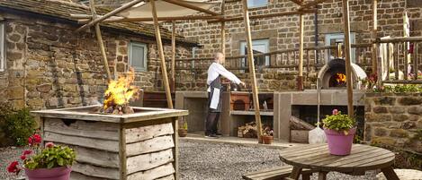 Ein Außenbereich mit Kochgelegenheit, einem Steinhaus, Holztischen und einem Pizzaofen.