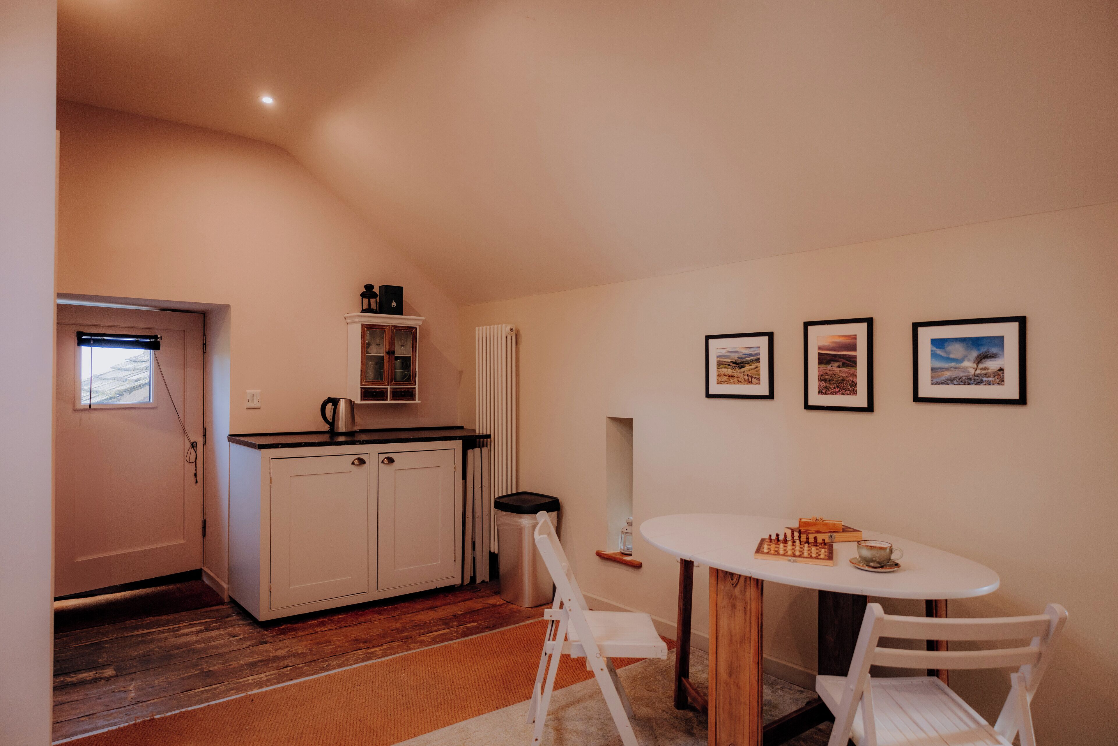 Une cuisine compacte avec des meubles blancs, un petit plan de travail et un radiateur. On y trouve une table à manger avec des chaises et un jeu d’échecs. Trois tableaux encadrés sont accrochés au mur.