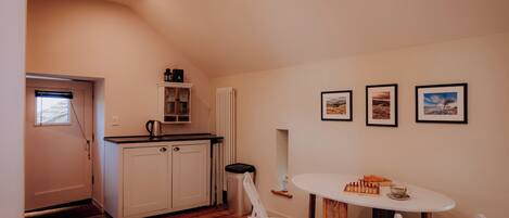 Une cuisine compacte avec des meubles blancs, un petit plan de travail et un radiateur. On y trouve une table à manger avec des chaises et un jeu d’échecs. Trois tableaux encadrés sont accrochés au mur.