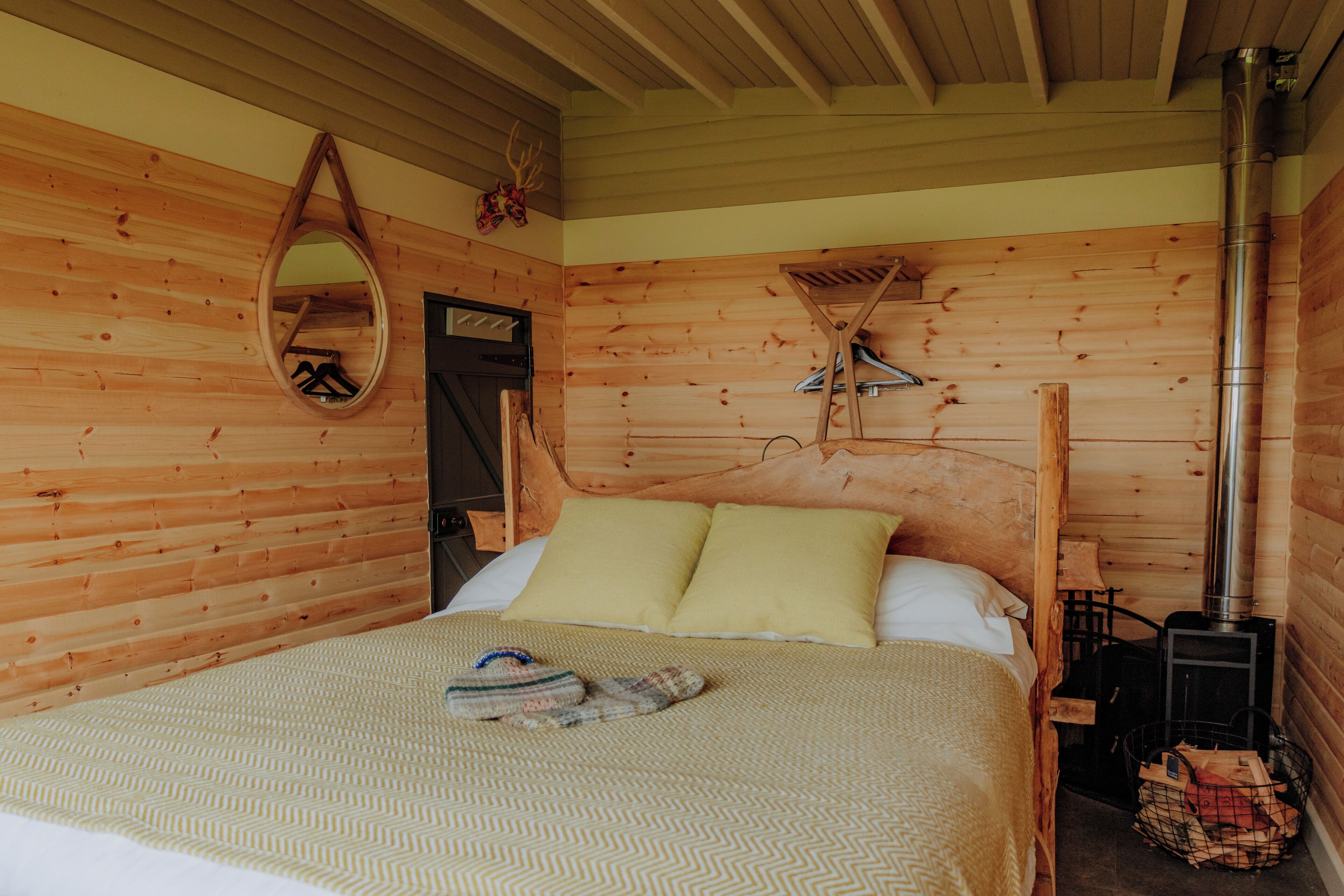 A wooden cabin bedroom with a bed, pillows, a hanging mirror, and a wood-burning stove.