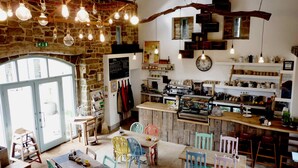A rustic cafe with exposed stone walls, wooden beams, and hanging light bulbs. There are colorful chairs and tables, a coffee machine, and shelves with kitchenware. - Swinton Bivouac (Ripon)