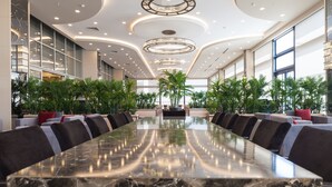 Uma sala de conferências ampla com uma longa mesa de mármore, cercada por sofás e cadeiras modernas, amplas janelas e luminárias decorativas no teto.
