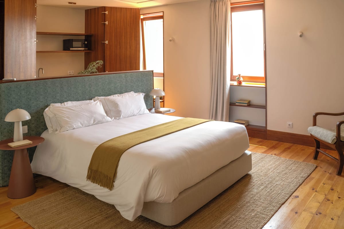 A neatly made bed with a headboard, bedside tables with lamps, a wooden floor, a chair, and a shelf with books and a small plant.