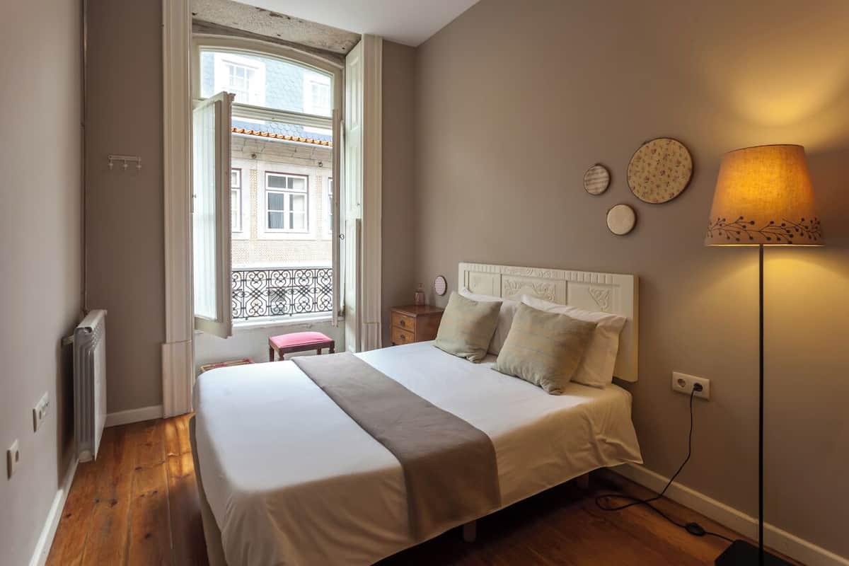 A bedroom with a bed, bedside table, lamp, and a window with a view of a building.