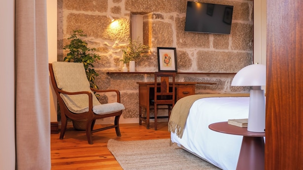 Un dormitorio con cama, una silla de madera con una toalla, un escritorio, una planta en maceta y un televisor instalado en una pared de piedra.
