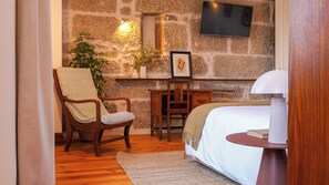 A bedroom with a bed, a wooden chair with a towel, a desk, a potted plant, and a TV mounted on a stone wall. - Being Porto Hostel (Porto)