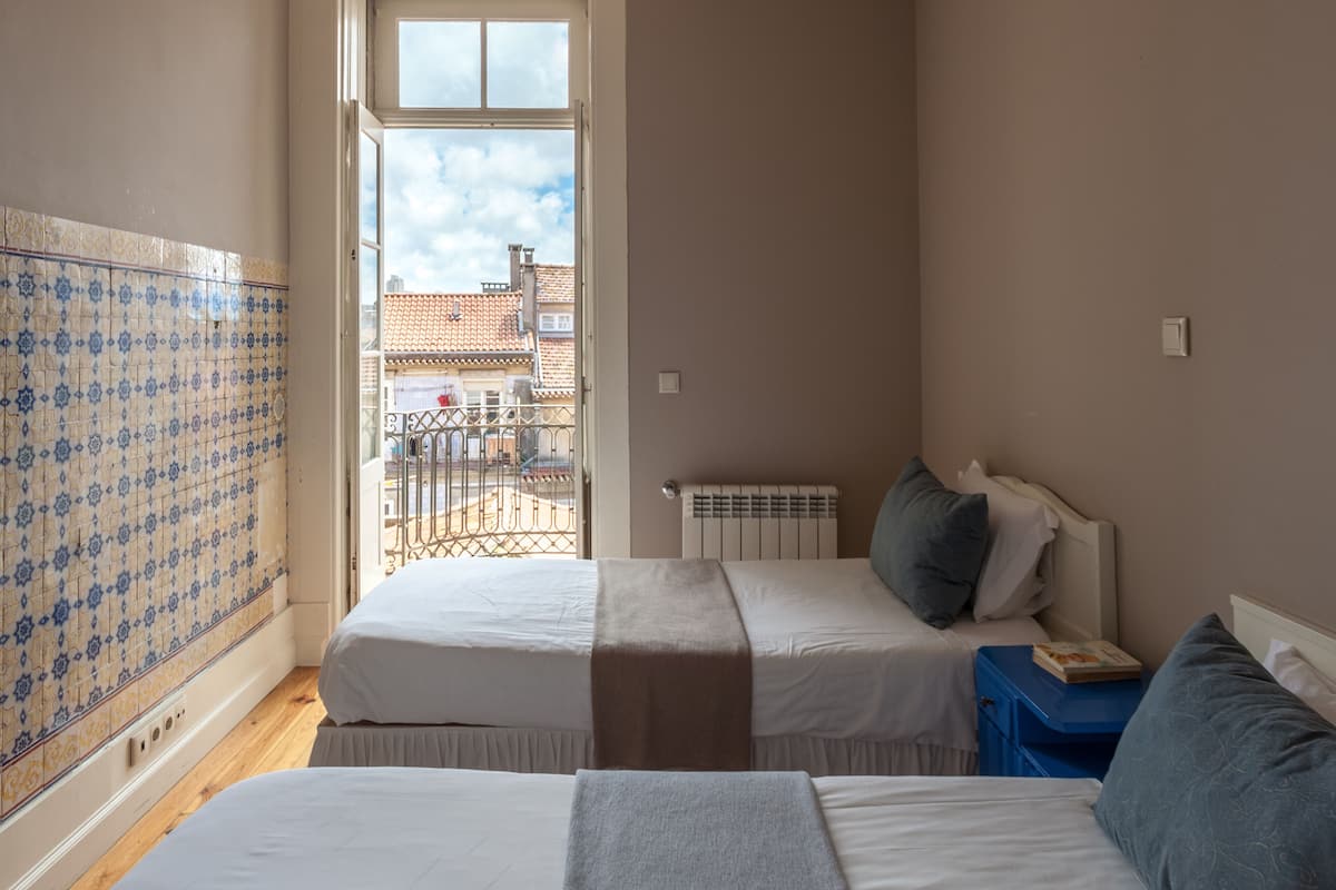 A bedroom with two beds, a radiator, and a window with a view of buildings.