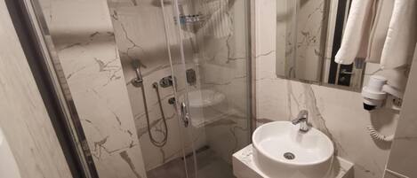 Un baño moderno con ducha envuelta en cristal, pared con azulejos de mármol, lavamanos blanco y secador de pelo montado en la pared.