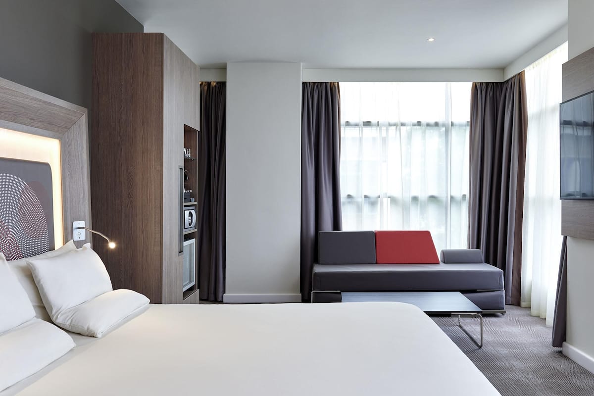 Hotellihuone, jossa on suuri sänky, punaisella koristetyynyllä varustettu sohva, työpöytä ja televisio.