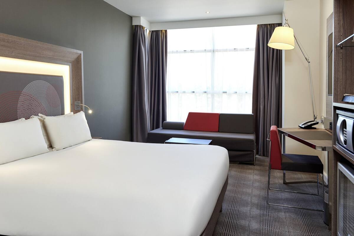 Une chambre d’hôtel avec un grand lit, un bureau avec une lampe, un canapé avec un coussin rouge et une grande fenêtre avec des rideaux.