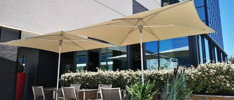An outdoor seating area with a large umbrella, stone wall, and modern building in the background.