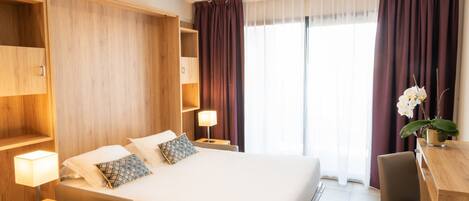 Habitación de hotel con una cama grande, cabecero de madera, mesitas de noche con lámparas, un escritorio con una silla y una ventana con cortinas.