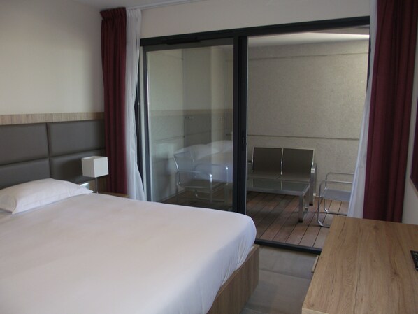 Habitación de hotel con cama, escritorio, silla y puerta corrediza de vidrio que da a un balcón.