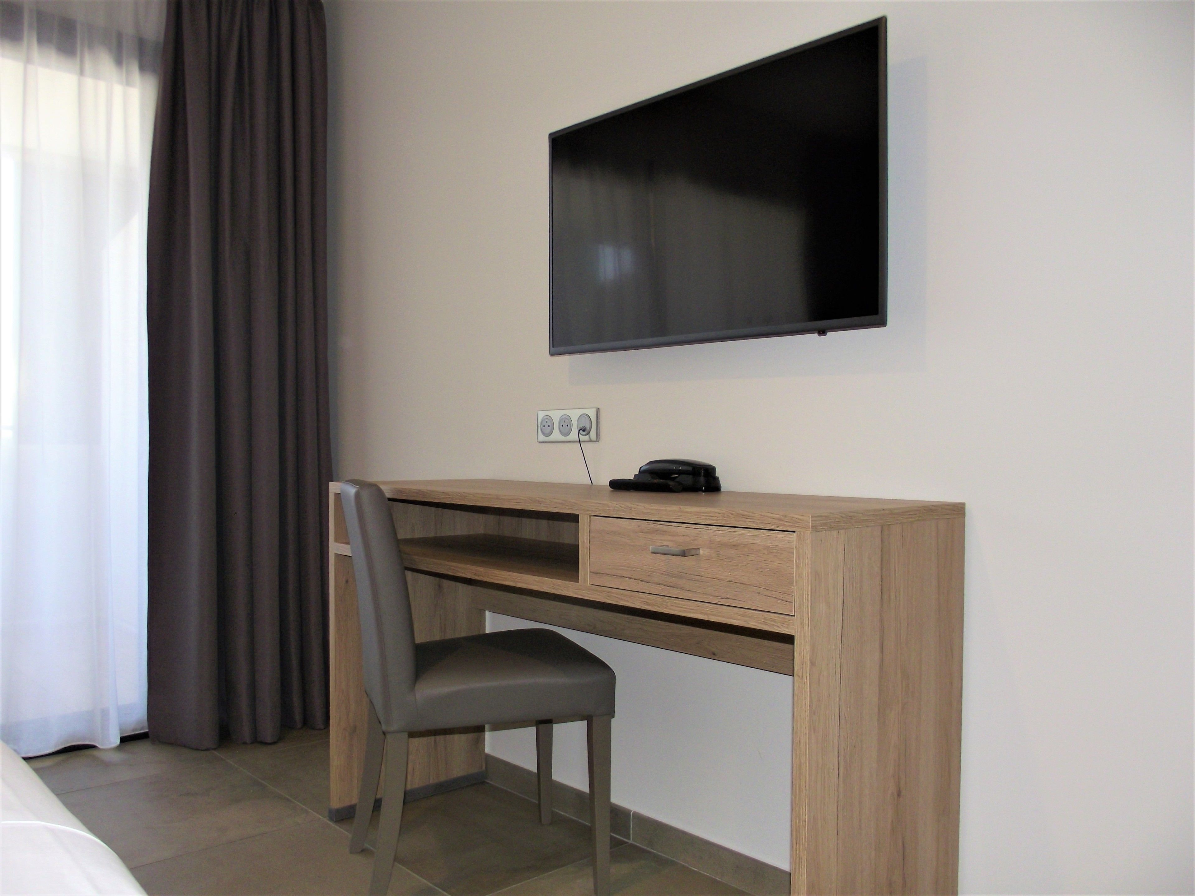 A hotel room with a wooden desk, a chair, a flat-screen TV mounted on the wall, and a power outlet.