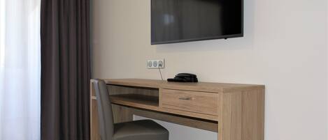 A hotel room with a wooden desk, a chair, a flat-screen TV mounted on the wall, and a power outlet.