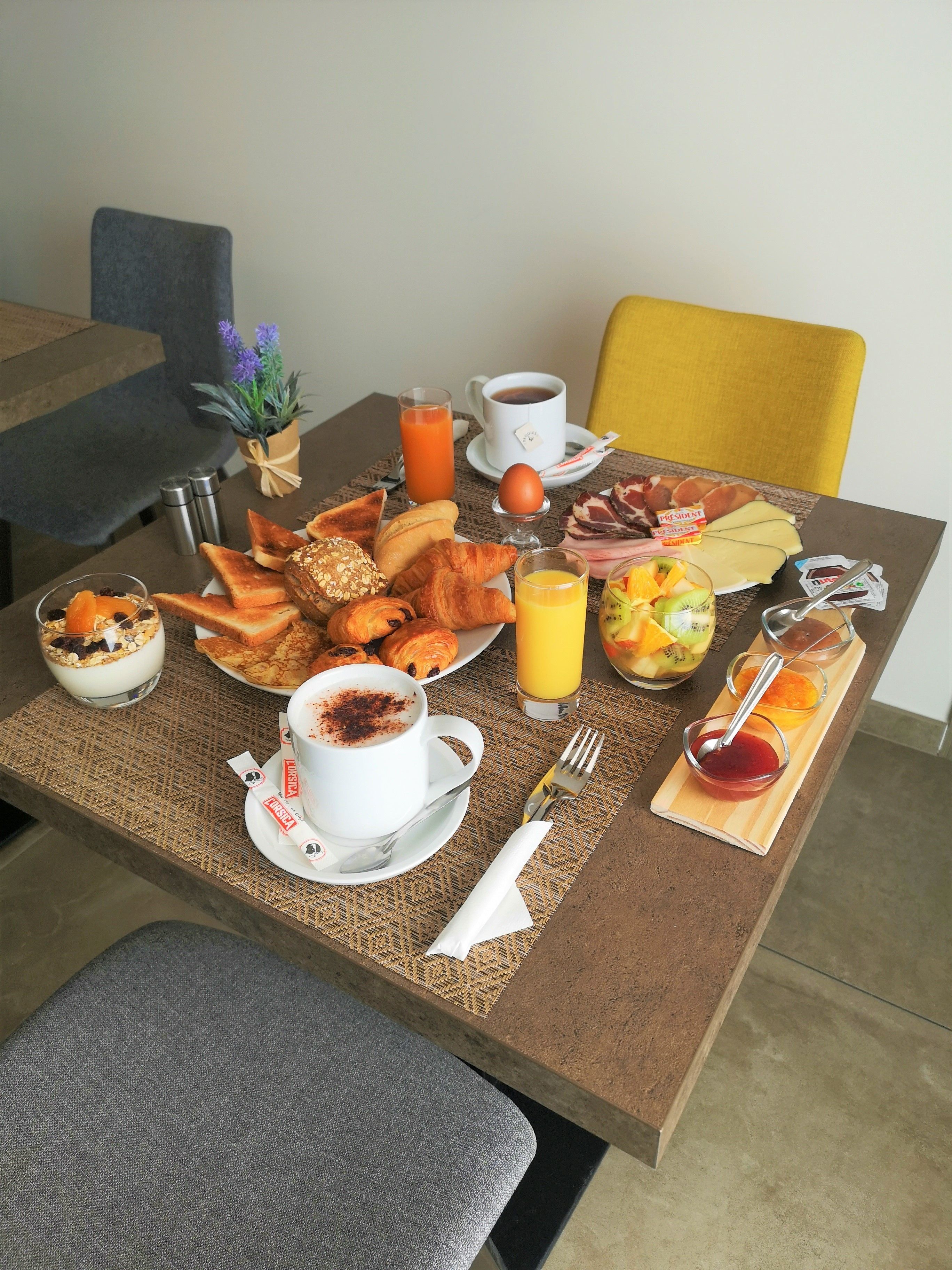 A breakfast table with croissants, toast, eggs, ham, cheese, and fruit. There are also cups of coffee and juice, and a small plant on the table.