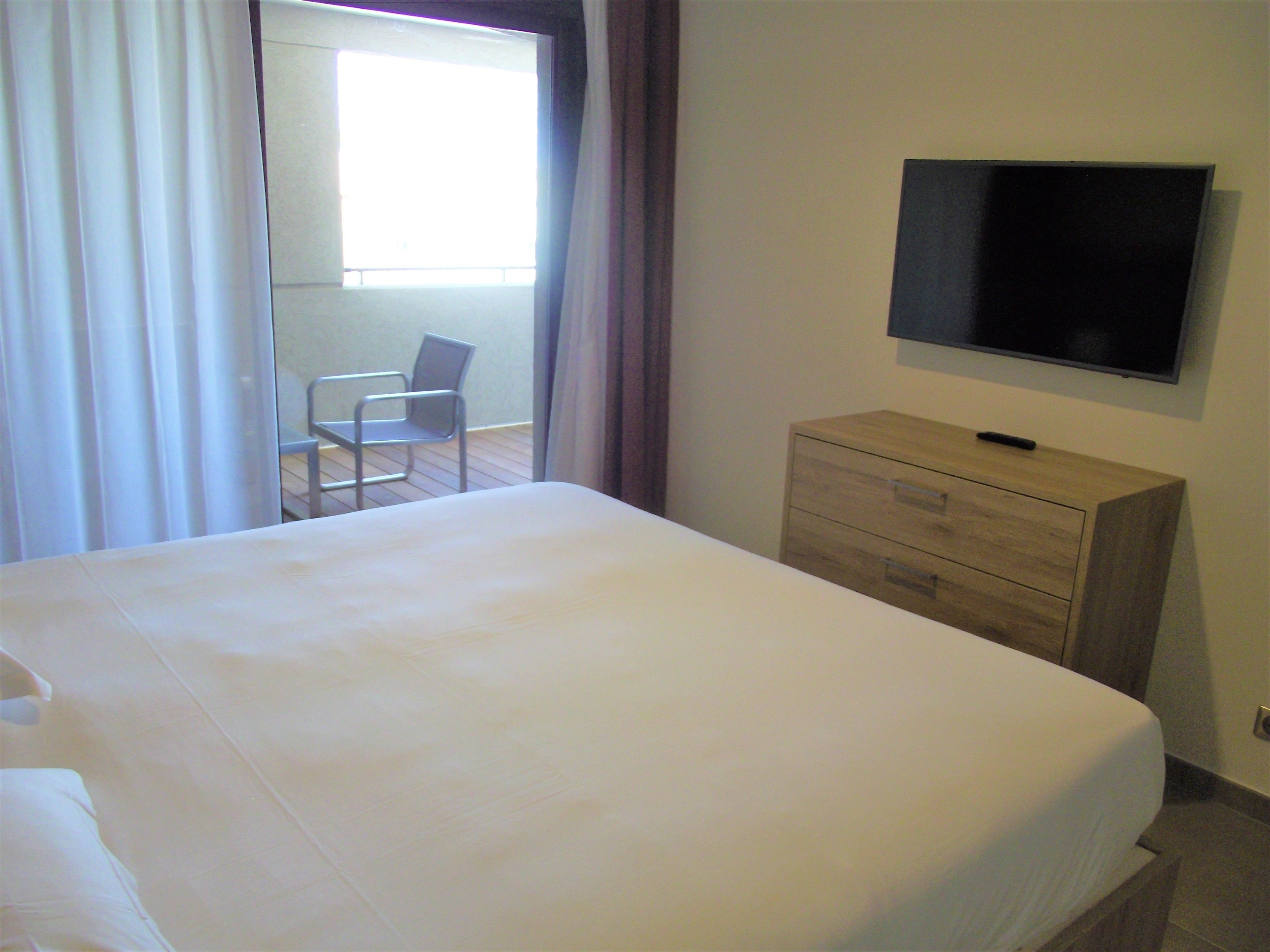 A hotel room with a bed, a wooden dresser, a television mounted on the wall, and a chair near a window.