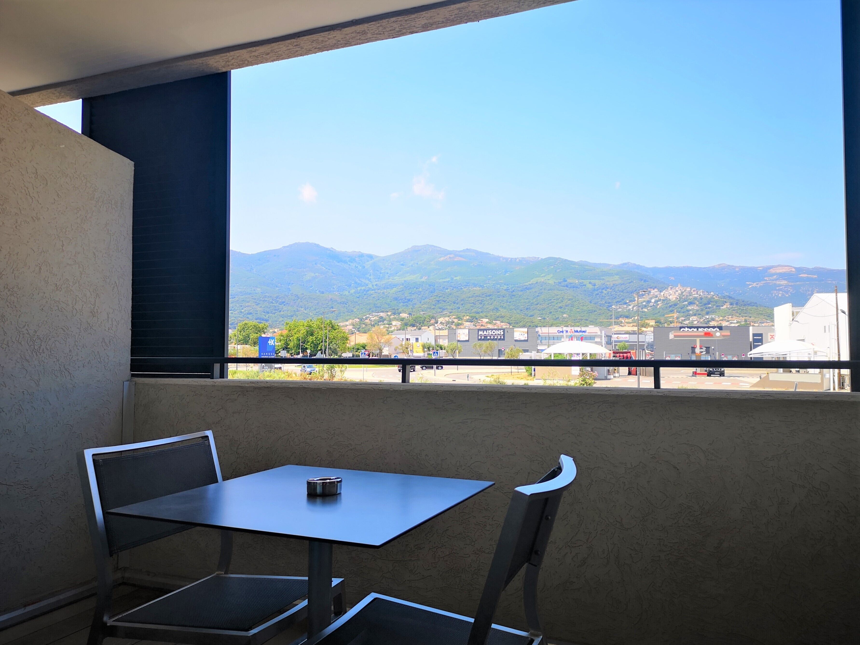A balcony with a table and chairs, overlooking a cityscape with mountains in the background.