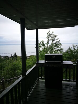 A covered outdoor area with a grill, wooden deck, and a view of the sea and greenery. - Auberge La Coudriere-Cool Hotel (Isle aux Coudres)