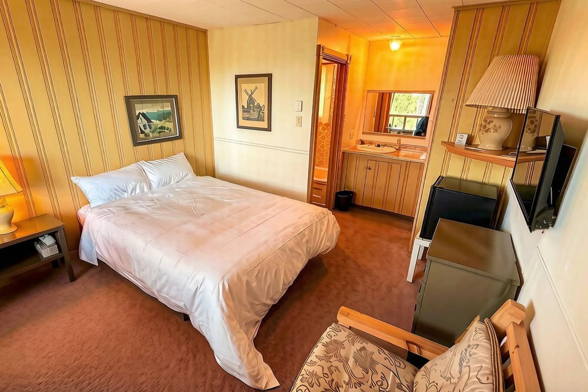 A hotel room with a bed, bedside table, lamp, and a small desk with a chair.