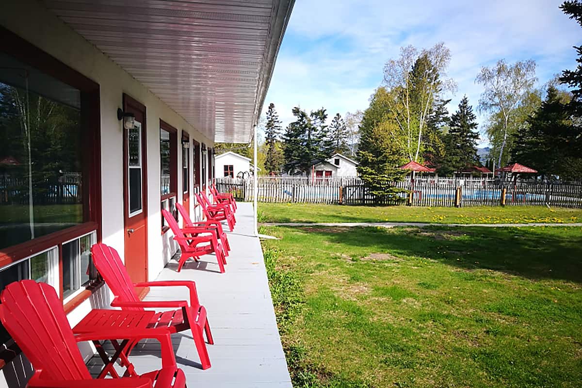 A covered porch with red chairs and tables, a white building with windows, and a fenced yard with trees and a small house in the background.