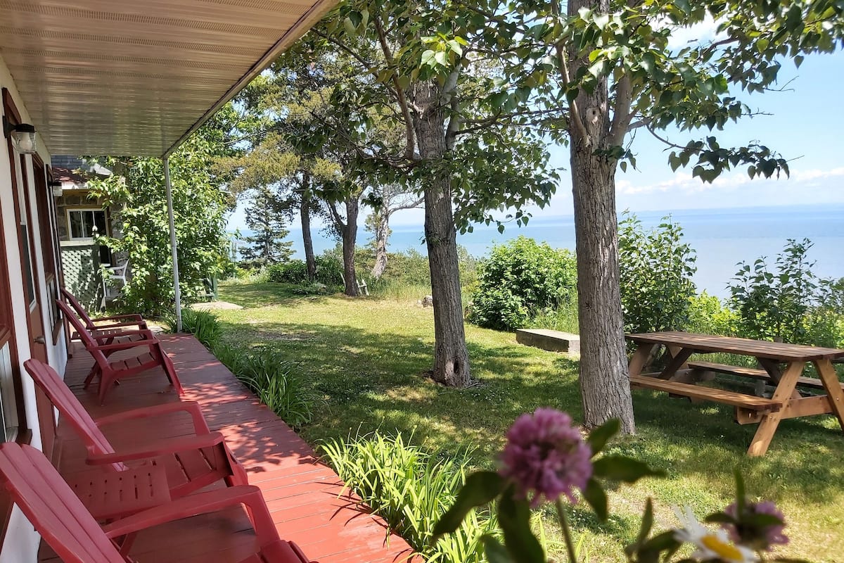 A covered outdoor seating area with red chairs and a picnic table, overlooking a grassy landscape and a body of water.