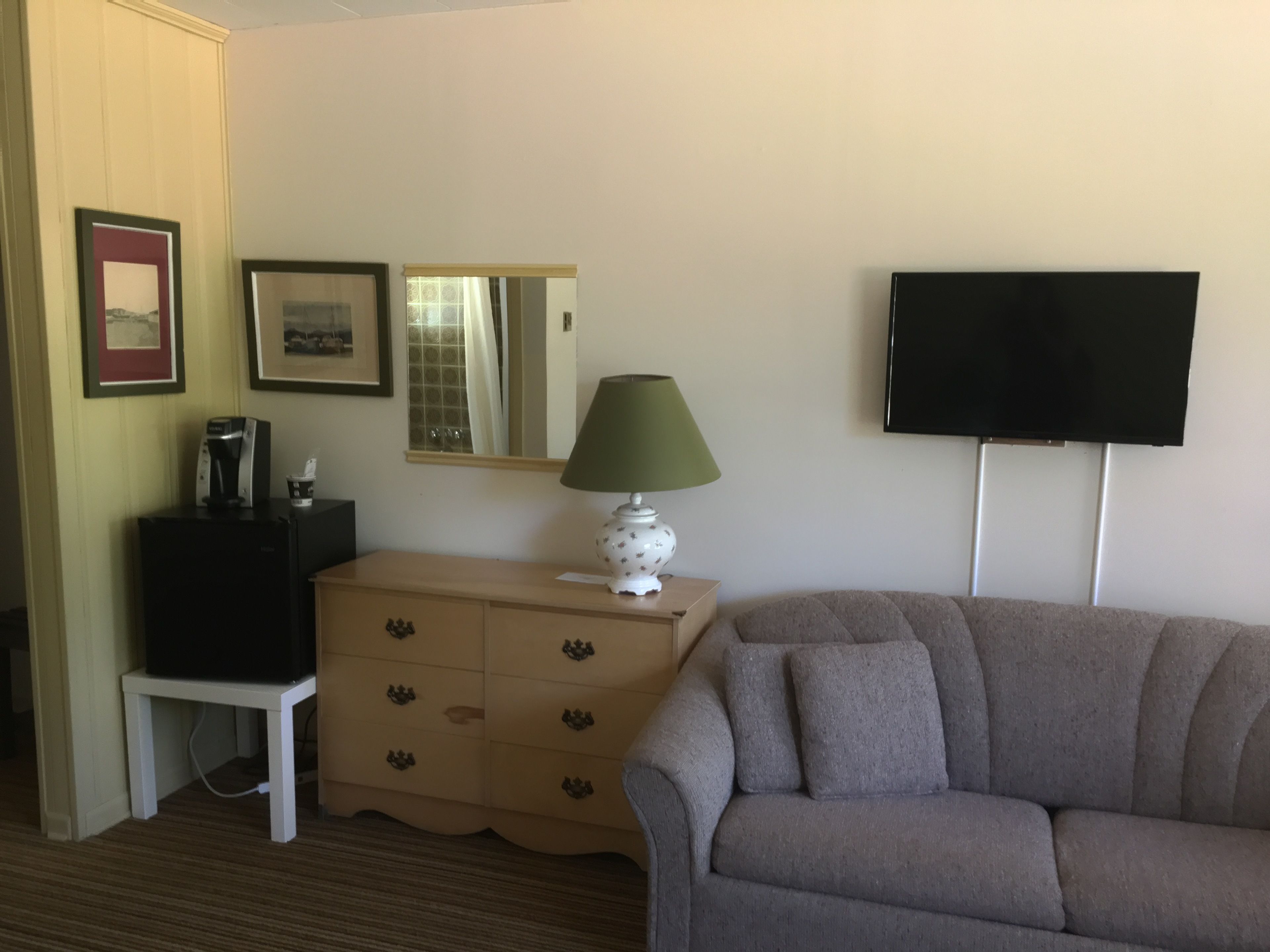 Ein Zimmer mit grauem Sofa, einem kleinen weißen Tisch mit Lampe, einer Kommode mit Schubladen, einem gerahmten Bild, einem Spiegel und einem Fernseher an der Wand.