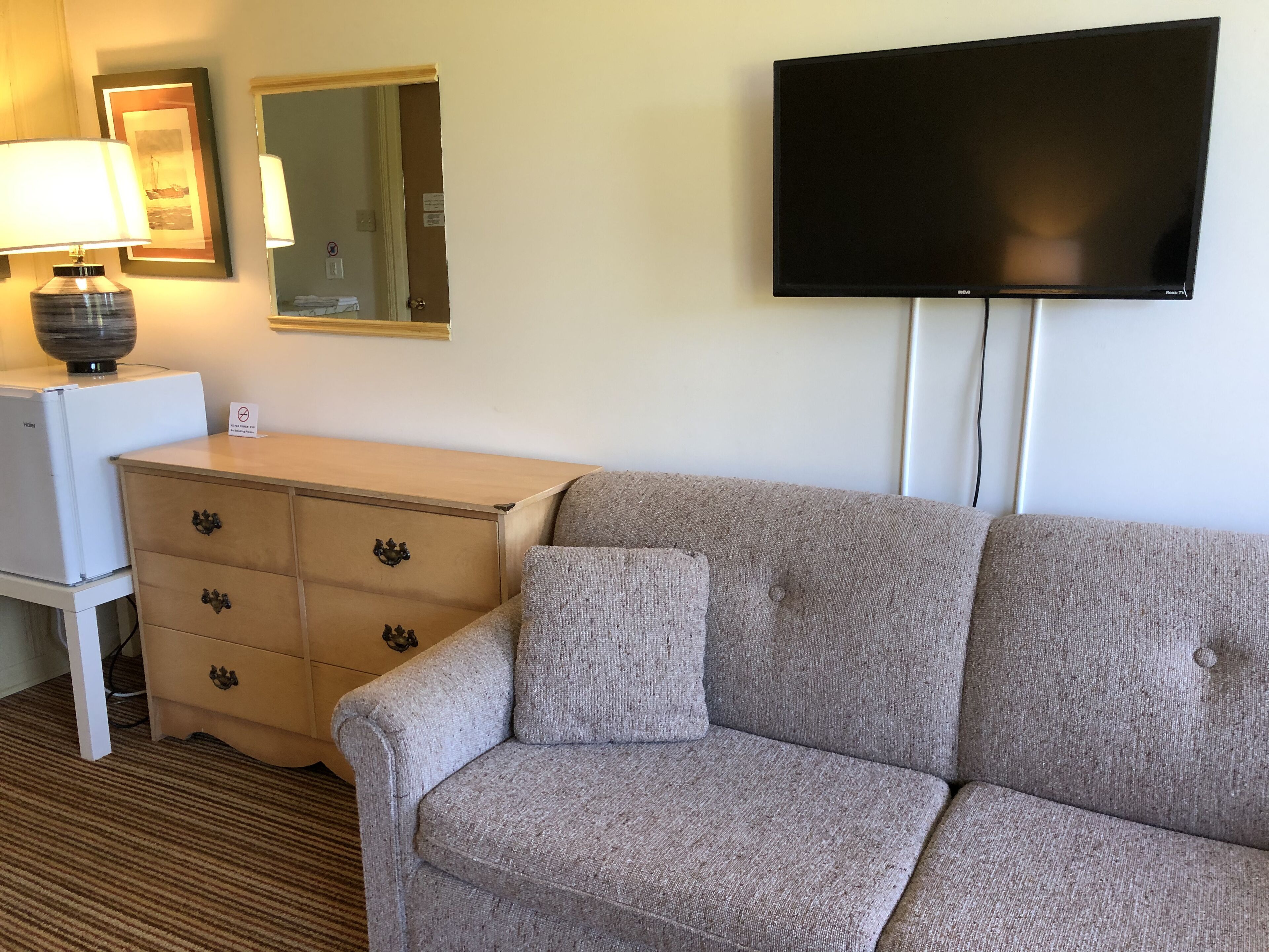 Una camera d'albergo con un divano color beige, un tavolino con una lampada, una televisione montata a parete e un frigorifero bianco.