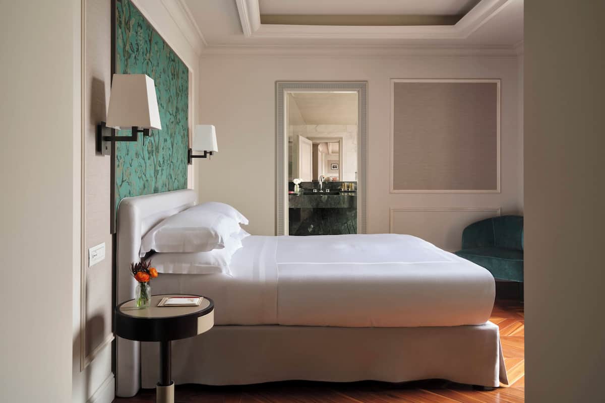 A neatly made bed in a hotel room with a small bedside table, a mirror, and a view into the bathroom.