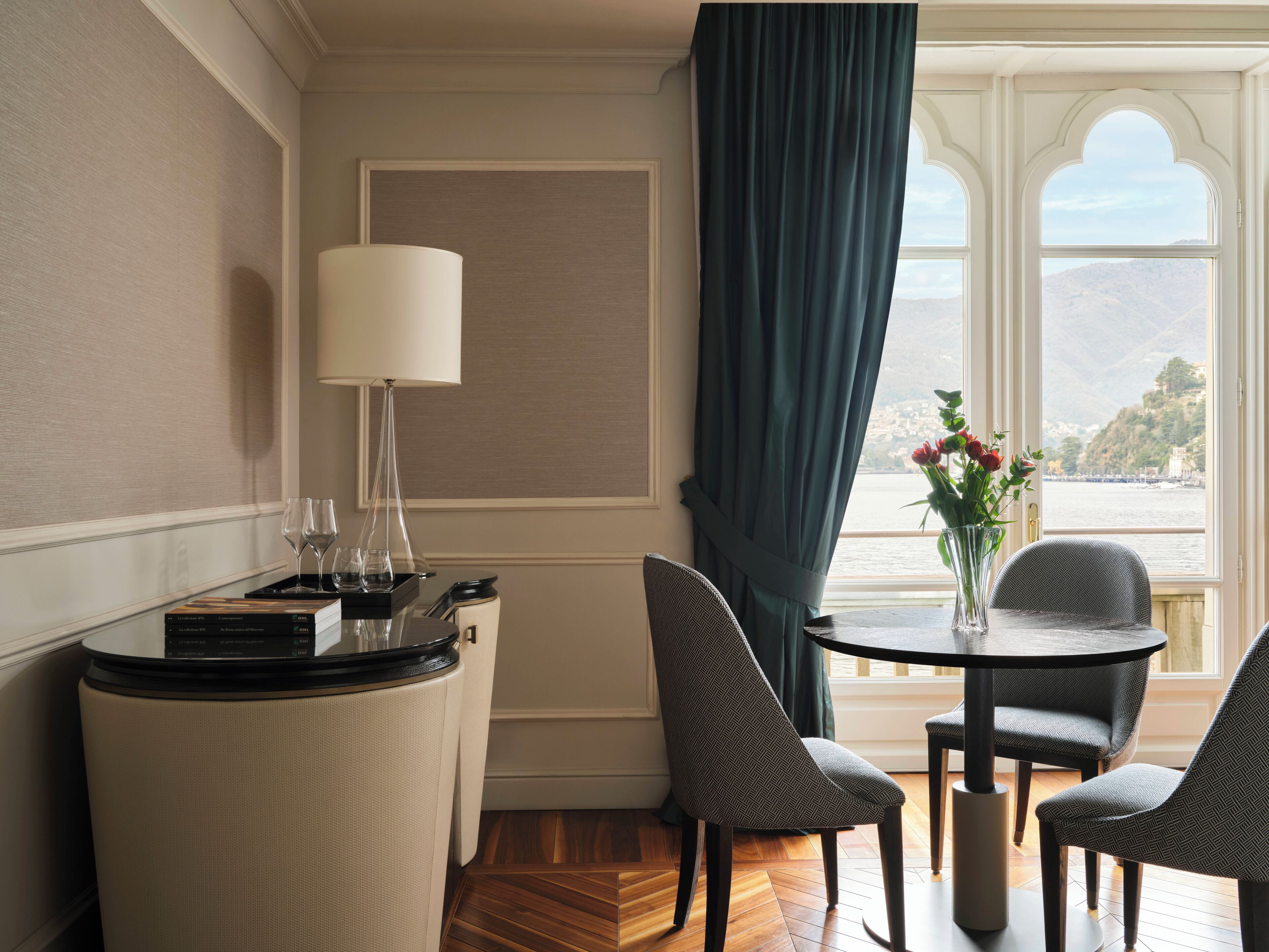 Une chambre d’hôtel dotée d’un coin repas comprenant une table ronde, deux chaises, une petite table d’appoint avec une lampe, et offrant une vue sur un plan d’eau grâce à une grande fenêtre munie d’un rideau.