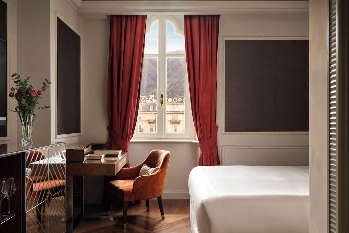 A hotel room with a bed, a desk with a chair, a window with curtains, and a view of buildings outside.