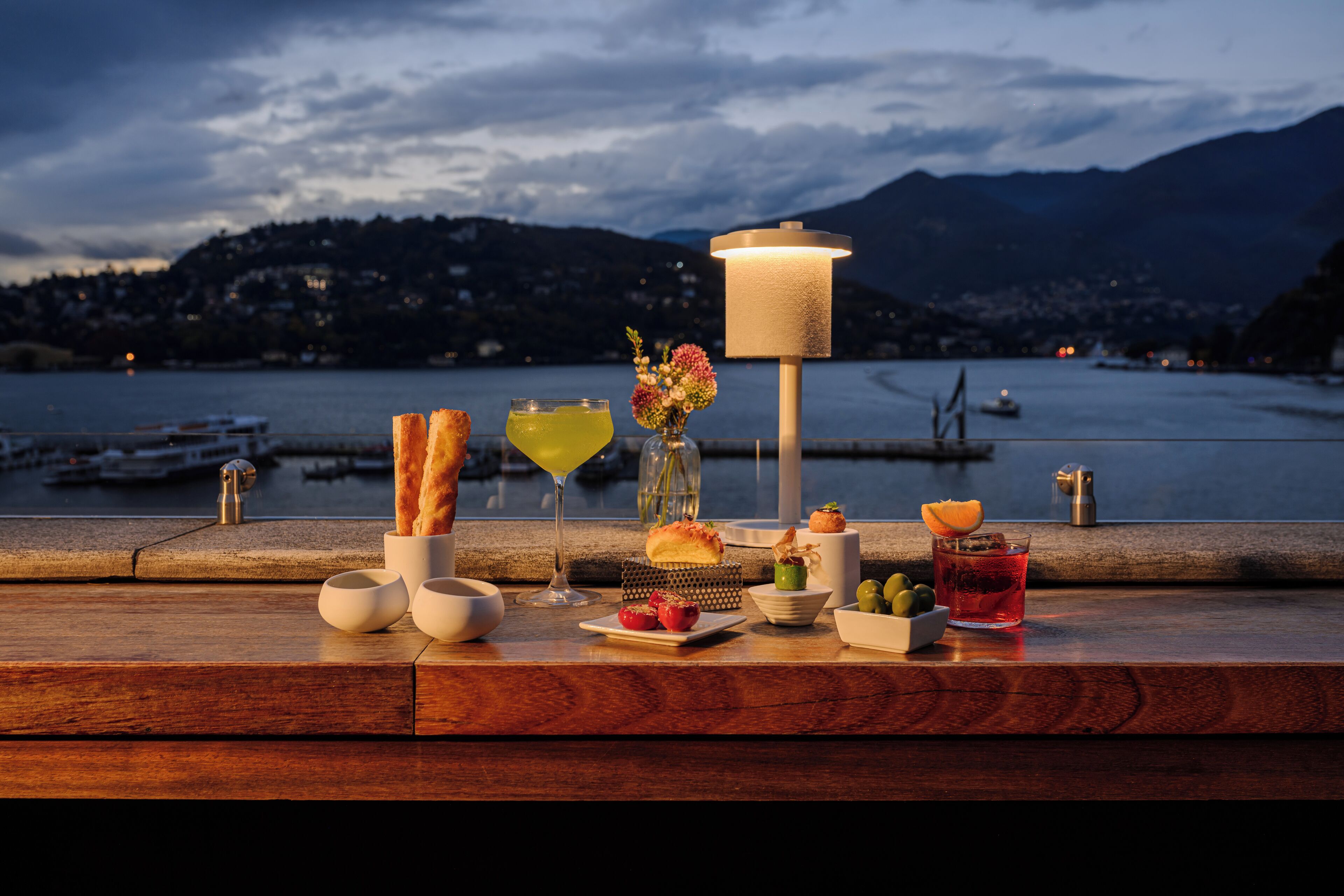 A table set with food and drinks, overlooking a body of water with mountains in the background.