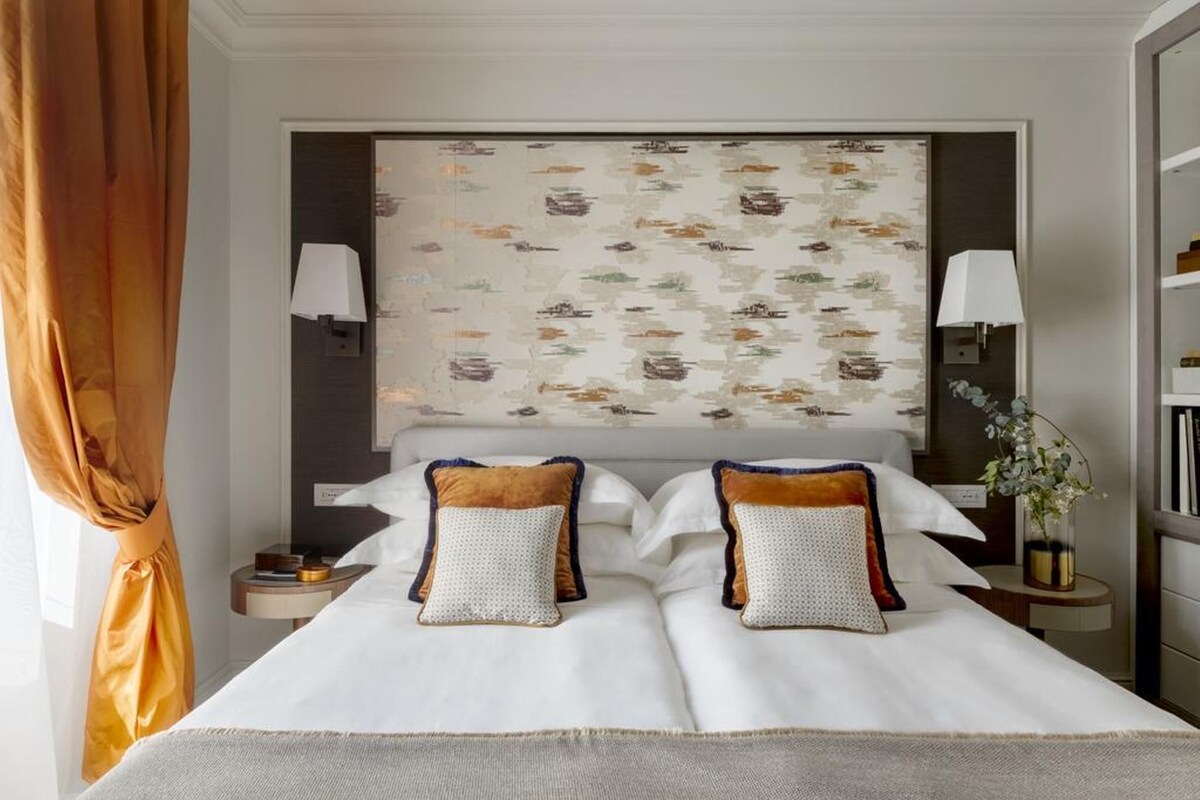 A hotel room with a bed, two bedside tables, a nightstand, a lamp, a wall-mounted artwork, and a curtain.