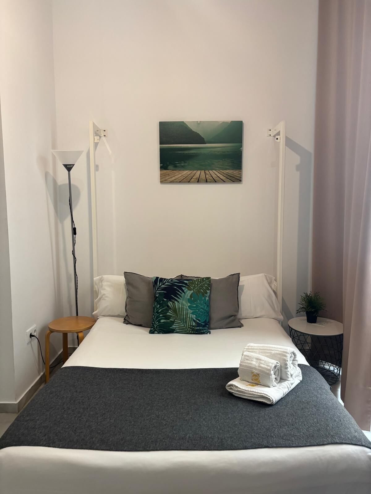 Una camera d'albergo con un letto, un quadro di un lago, una lampada da terra, un tavolino e una pianta in vaso.