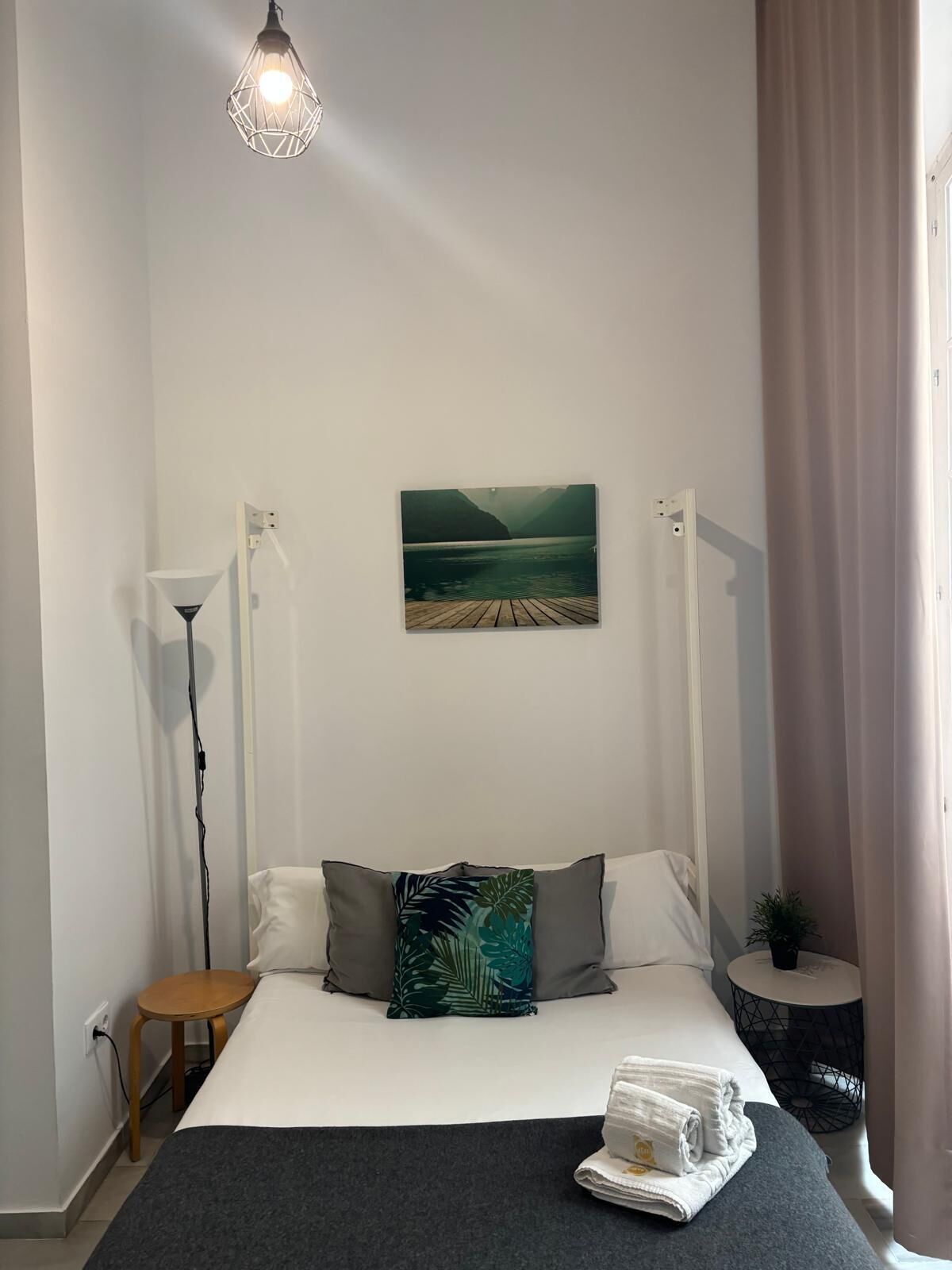 A bedroom with a bed, a painting of a lake, a lamp, and a small table.