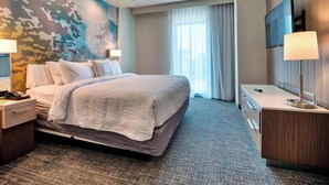 Habitación de hotel con una cama grande, un escritorio con lámpara, un televisor montado en la pared y un amplio ventanal con cortinas.