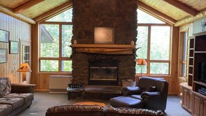 A cozy living room with a stone fireplace, wooden ceiling beams, large windows, and a television. - Gunflint Lodge & Outfitters (Grand Marais)