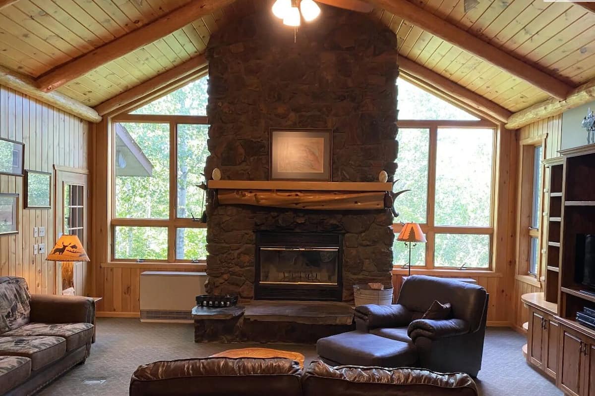 A cozy living room with a stone fireplace, wooden ceiling beams, large windows, and a television.