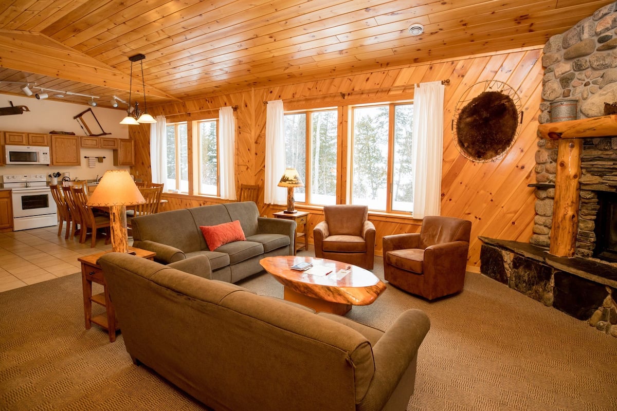 A cozy cabin interior with a wooden ceiling, a stone fireplace, a dining area with a table and chairs, and a living room with a sofa and armchairs.