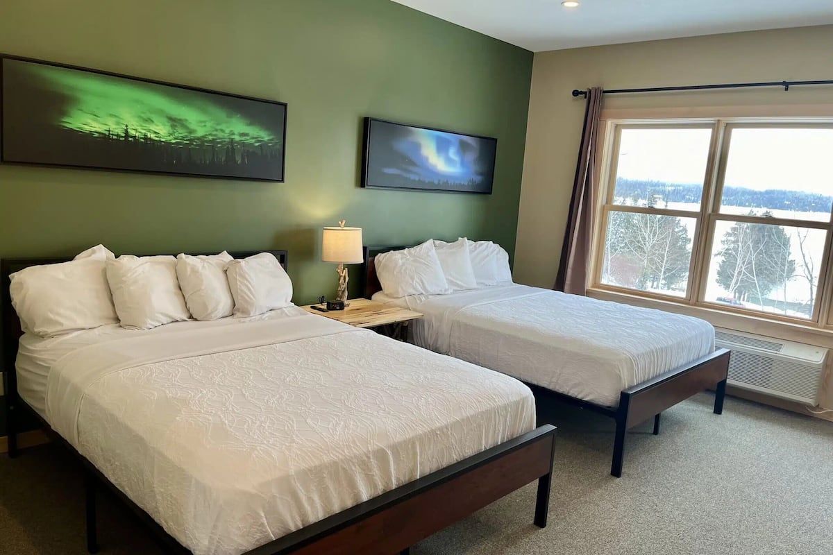 A hotel room with two beds, a nightstand with a lamp, and a large window offering a view of trees and a snowy landscape.